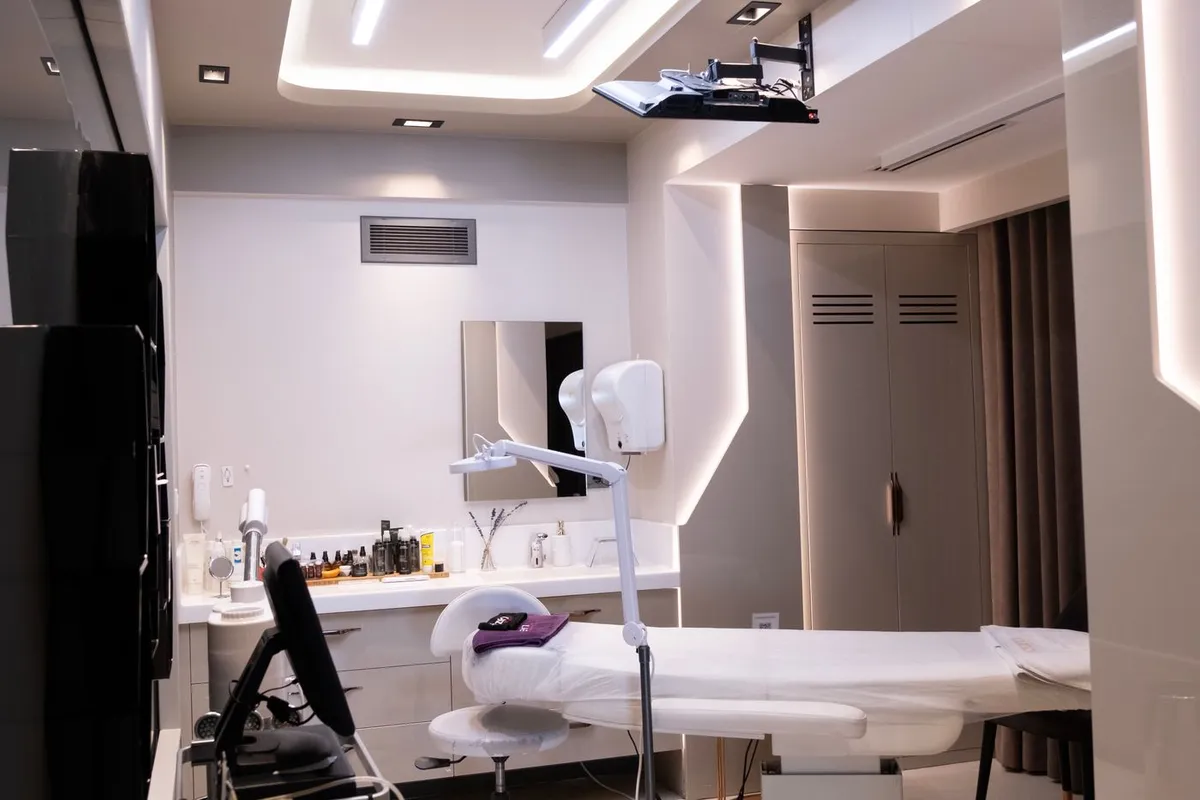 A treatment room in an aesthetics clinic: treatment couch, magnifying lamp and a product shelf under warm lighting.