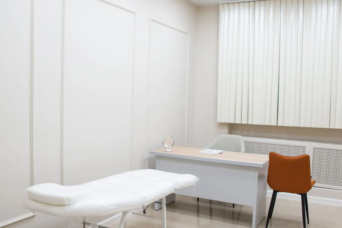 A consulting room set up for a clinic session: examination couch, desk and chair against a window.
